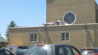 A priest holding mass on the roof of a church as cars line up in the parking lot to listen to the service.