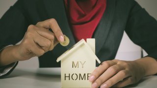 mujer toma una moneda en una mano y en la otra sostiene una alcancía con forma de casa