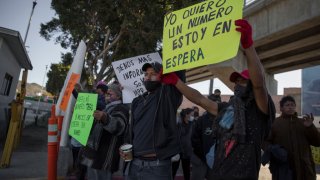 Migrantes en la frontera entre San Diego y Tijuana con letrero yo quiero un número estoy en espera