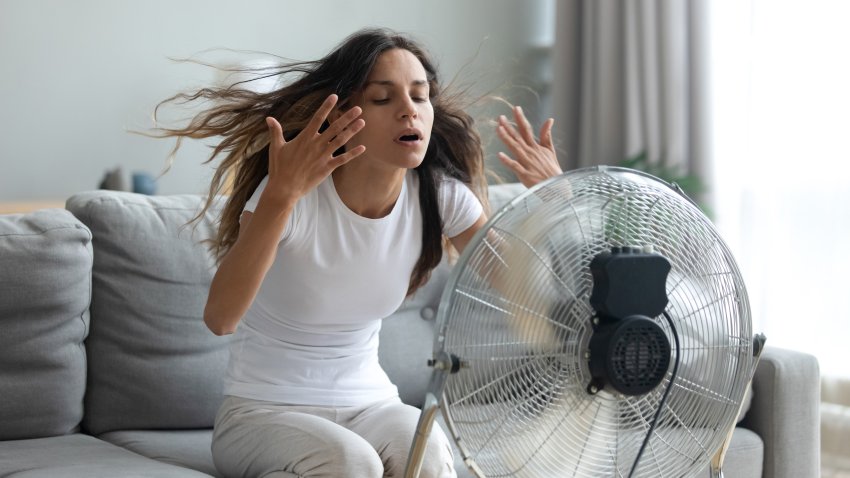 In living room without air-conditioner tired from summer heat young woman turned on floor ventilator waving her hands to cool herself, female sitting on couch suffers from unbearable too hot weather