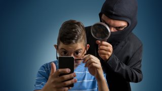 Foto concepto que muestra a joven con celular y alguien enmascarado que espía lo que hace con el dispositivo.