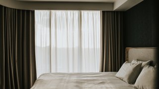A calm and serene scene within an apartment or hotel room. Low sun shines through a large window, diffused by net curtains. A neatly made bed is visible in the foreground.