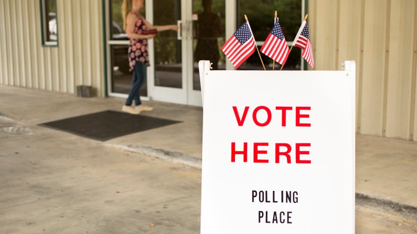 ‘Vote Here, Polling Place’ sign outside of a local, public polling location in USA.  American flags top the sign.  One woman enters the building.  The USA elections are held in November each year.