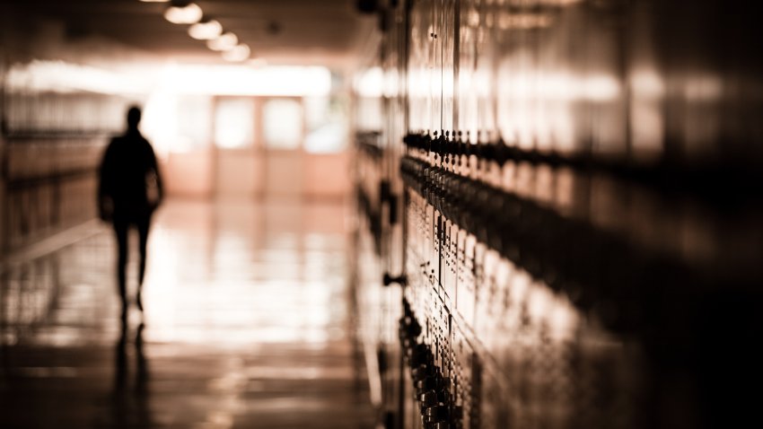 A high school student walks down a dark hallway in a public high school, silhouetted by daylight spilling in and reflecting off of the floor and lockers.