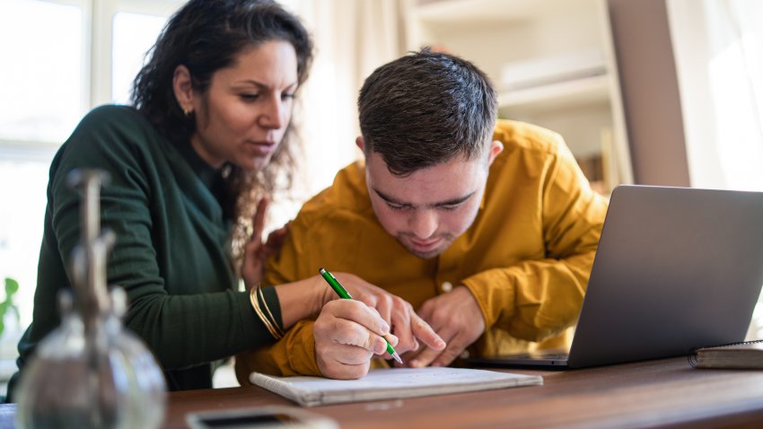 Mid adult woman helping young man with Down syndrome with learning and doing homework online on laptop.