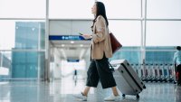 Young Asian woman carrying suitcase and holding smartphone on hand, walking in airport terminal. Ready to travel. Travel and vacation concept. Business person on business trip