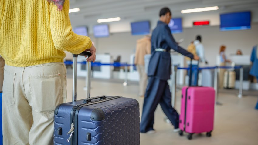 A traveler stands at an airport check-in counter with a yellow sweater and rolling suitcase, ready for a trip. The scene reflects anticipation and movement.
