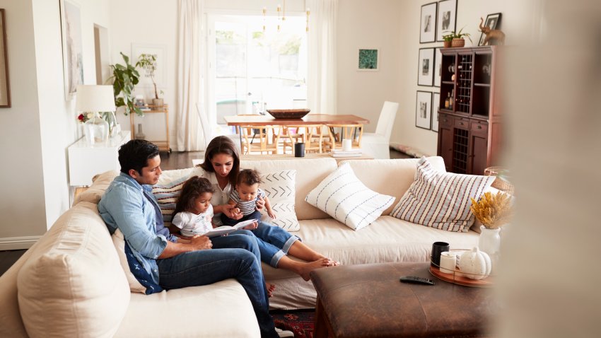 Young Hispanic family sitting on sofa reading a book together in the living room, seen from doorway
