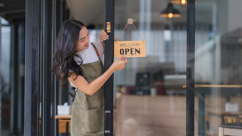 Asian woman entrepreneur hanging an open sign on glass door, welcoming customers to her small business coffee shop