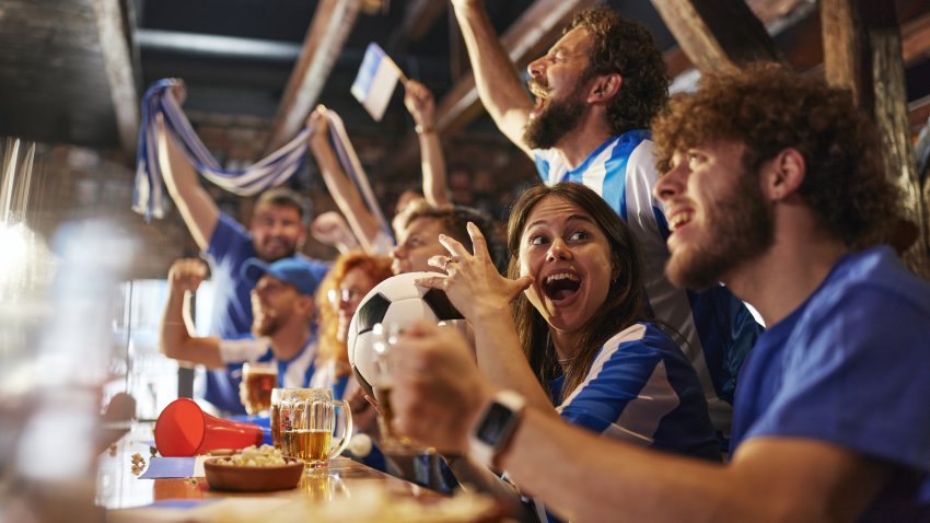 Group of cheerful sports fans cheering while watching a soccer match on TV in a bar. Focus is on woman with ball.